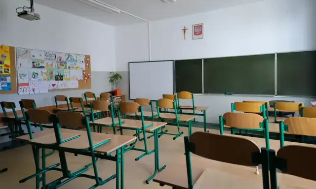 Pusta klasa szkolna z rzędami ławek i krzeseł. Na ścianie tablica, projektor i dziecięce rysunki. Wygląda na to, że **odwołane zajęcia w szkołach** sprawiły, że nikt dziś nie przyszedł.