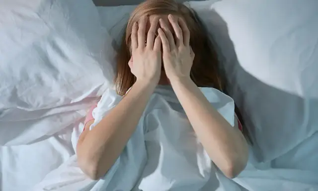 Nastolatka w łóżku, zakrywająca twarz dłońmi, symbolizuje bezsenność u nastolatków i jej przyczyny.