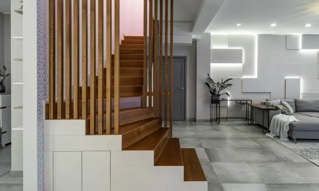 Nowoczesna, wysoka klatka schodowa aranżacje z drewnianymi stopniami i ażurową balustradą, wkomponowana w minimalistyczne wnętrze z szarą sofą i geometryczną ścianą.
