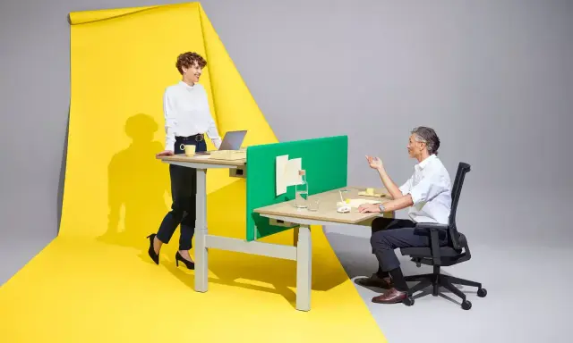 Femme en position debout statique, souriante, devant un bureau. Homme assis à un bureau, discutant. Fond jaune et gris.