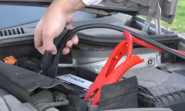 Mano conectando pinzas rojas a la batería de un coche para arrancar con pinzas.