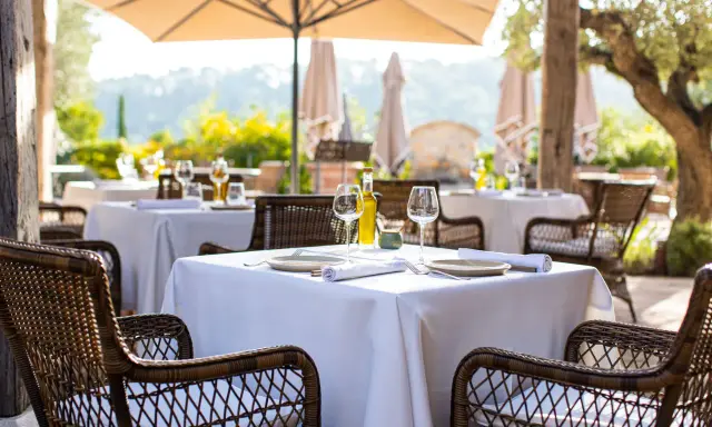 Tables dressées en terrasse, sous un parasol, invitant à savourer une cuisine du terroir authentique. Vins, verres et couverts attendent les convives.