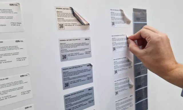 Ręka zrywa etykietę z folii, która jest surowcem dla drukarni. Widać różne rodzaje folii, m.in. srebrną matową i białą błyszczącą.