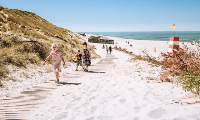 Familj på väg längs en träspång över vit sand i Danmark. En rödvit fyr och bunkrar syns i bakgrunden.