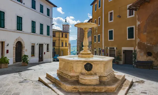 Fontana in pietra nella Valdichiana Senese, circondata da edifici storici con persiane verdi e un cielo azzurro con nuvole bianche.