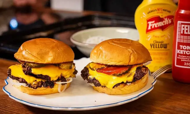 Två saftiga hamburgare med smält ost, pickles och lök på en tallrik. Bredvid står flaskor med senap och ketchup.