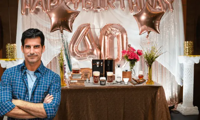 Mężczyzna w kratę stoi przed stołem z prezentami i balonami z napisem "Happy Birthday 40".
