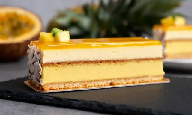 Un délice tropical : un entremets aux couches crémeuses, surmonté d'un glacage miroir jaune éclatant, décoré de fruits et de noix de coco râpées.