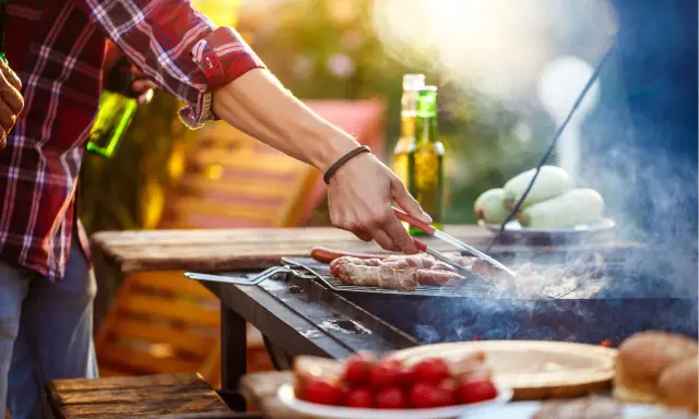 Hombre asa salchichas envueltas en tocino, ideal para saber qué llevar a una barbacoa para sorprender. Hay cervezas y tomates en la mesa.