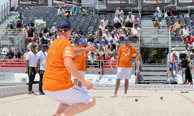 Joueur de pétanque concentré, cherchant à faire le vide dans sa tête avant de lancer la boule. Le public est installé dans les gradins derrière lui.