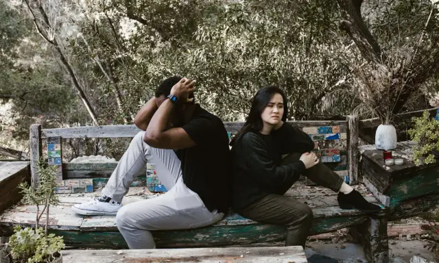 Pareja sentada de espaldas en un banco, con expresiones de angustia. El hombre se agarra la cabeza, ella mira al vacío. Un **vínculo traumático** parece separarlos en este entorno natural.