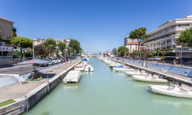 La darsena di Riccione, con barche ormeggiate lungo il canale, palazzi e alberi verdi sotto un cielo azzurro.