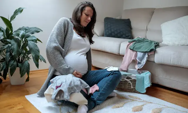Schwangere Frau sitzt auf dem Boden, hält ihren Bauch und scheint Braxton Hicks Kontraktionen zu spüren, während sie Babykleidung sortiert.