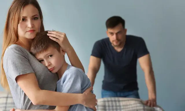 Smutna kobieta tuli syna, podczas gdy partner patrzy z dystansu. Trudno jej zaakceptować dziecko partnera.