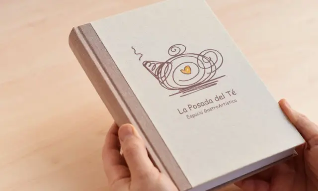 Manos sostienen un libro con tapa de tela, con un diseño de tetera y el texto "La Posada del Té". Una idea para como encuadernar un trabajo de forma original.