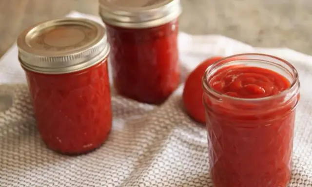Trois bocaux de ketchup maison, un rouge vif, reposent sur une serviette à motifs. Un tomate entière est visible.
