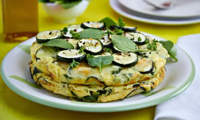 Deliciosa tortilla de calabacín apilada, adornada con rodajas de calabacín, espinacas y perejil fresco.
