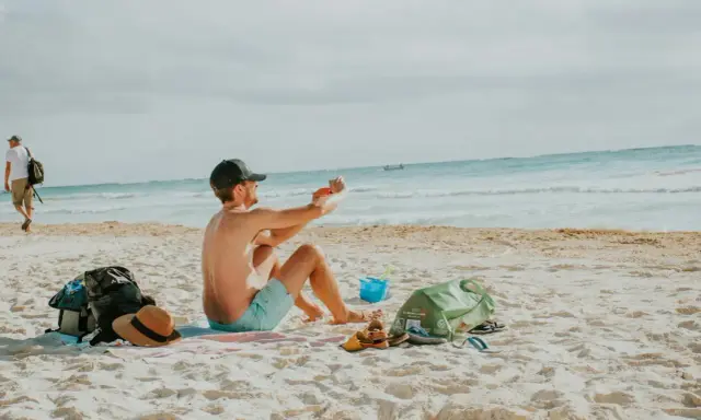Mężczyzna robi zdjęcie telefonem na plaży, marząc o najkrótszych lotach z Polski na wakacje. Obok leżą torba, kapelusz i sandały.