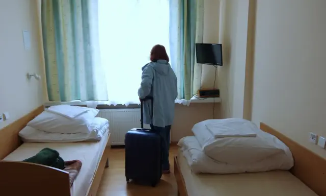 Kobieta z walizką przy oknie, gotowa by dowiedzieć się, jak wyjechać do sanatorium. Pokój z dwoma łóżkami.