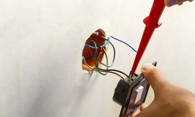 Ręce montują włącznik światła, podłączając przewody. Widać instalację elektryczną i narzędzie.