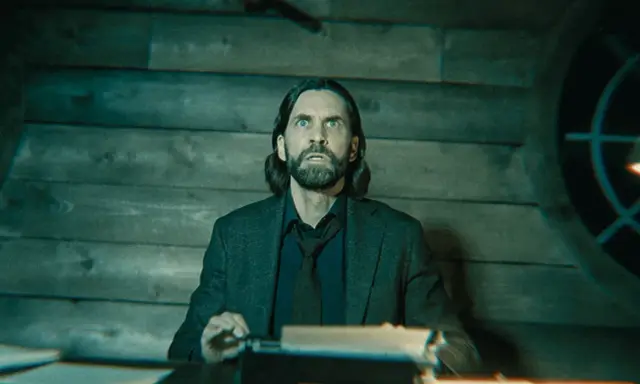 Alan Wake 2: un hombre con barba y pelo largo, con expresión de sorpresa, sentado frente a una máquina de escribir en una habitación de madera.