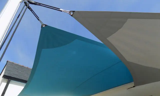 Comment installer un voile d'ombrage sur terrasse : deux voiles, bleu et gris, fixés par des cordages et des poulies à un mât et à la maison.