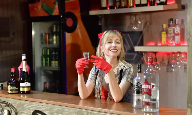 Uśmiechnięta barmanka w czerwonych rękawiczkach trzyma kieliszek. Na ladzie stoi duża butelka wódki, idealna na pół litra na dwóch.