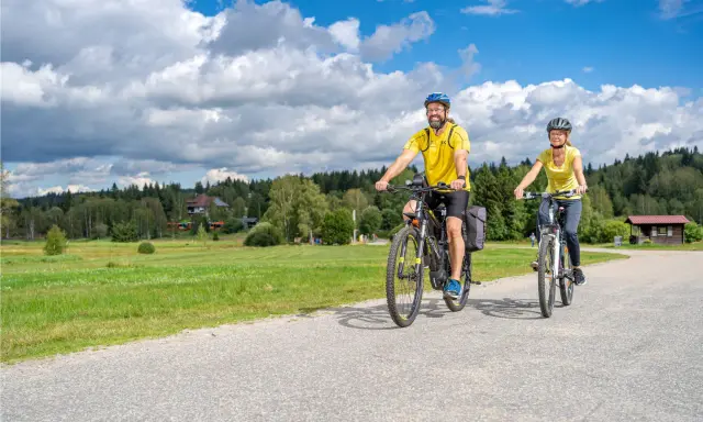 Glückliches Paar auf E-Bikes auf einer Tour durch den Bayerischen Wald. Die Sonne scheint, der Himmel ist blau mit weißen Wolken.