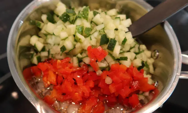 Finhackad gurka och röd paprika i en kastrull, redo för ett bostongurka recept. En sked vilar i blandningen.