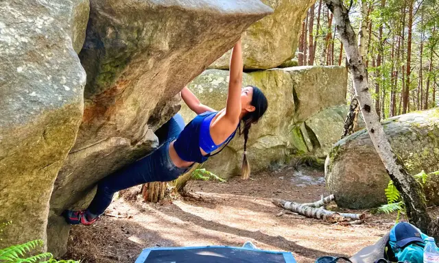 Kobieta wspina się po skale w lesie, doświadczając emocji związanych z boulderingiem w Fontainebleau.