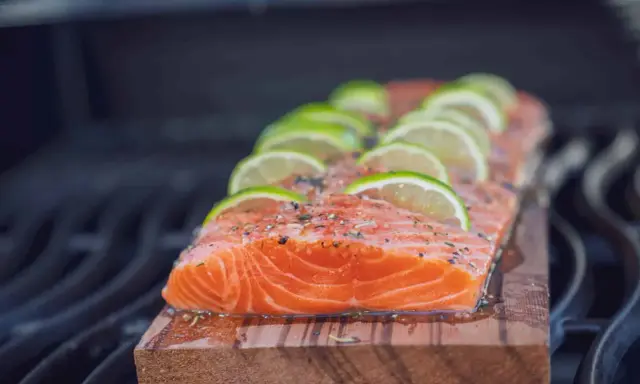 Ein saftiges Lachsfilet mit Limettenscheiben und Kräutern wird auf einem Holzbrett gegrillt.