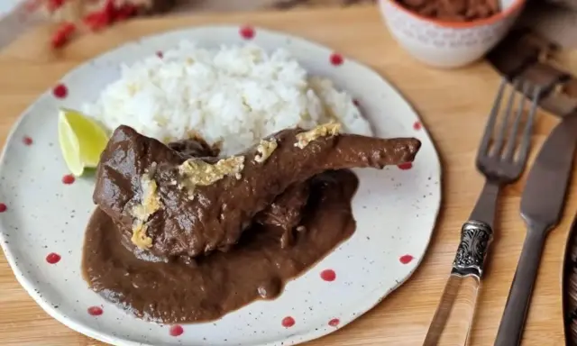 Un delicioso conejo guisado en mole oscuro, servido con arroz blanco y una rodaja de limón.