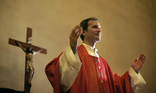Kapłan w czerwonej szacie z podniesionymi rękami, obok krzyża z Jezusem. Wiernie przestrzega zasad prawa kanonicznego.
