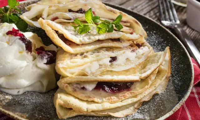 Stos naleśników bez mleka z dżemem wiśniowym i bitą śmietaną, ozdobiony miętą i posypany cukrem pudrem.