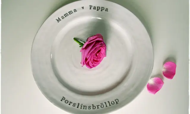 En vit tallrik med texten "Mamma ♡ Pappa" och "Porslinsbröllop". En rosa ros ligger i mitten. Ett vackert minne för de som varit gifta i 20 år.