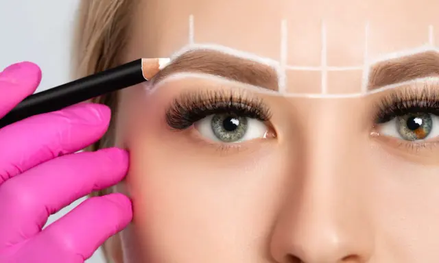 Main d'une esthéticienne en gant rose dessinant la forme sourcil visage rond avec un crayon blanc.