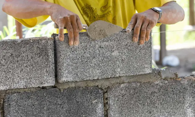 Budowa fundamentu z bloczków betonowych. Ręce murarza układają kolejny bloczek, używając kielni z zaprawą.