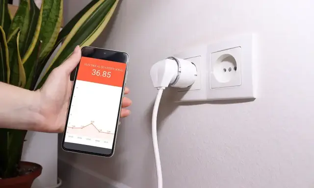 Smart wtyczka w gniazdku i smartfon z wykresem pokazującym monitoring zużycia energii.