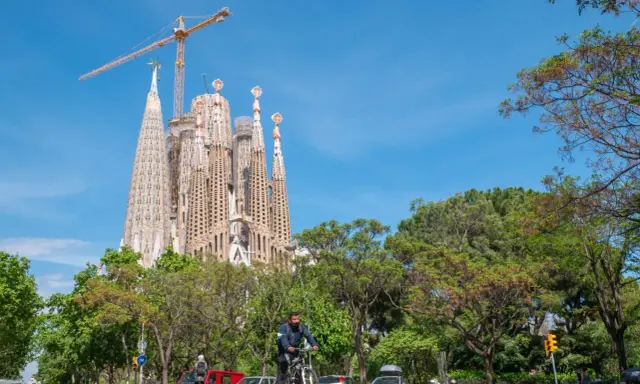 Sagrada Familia w Barcelonie, wciąż w budowie, góruje nad zielonym parkiem. Na ulicy widać rowerzystę i samochody.