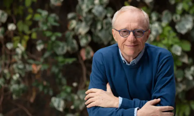 David Grossman, con occhiali blu e maglione scuro, sorride con le braccia incrociate su sfondo verde.