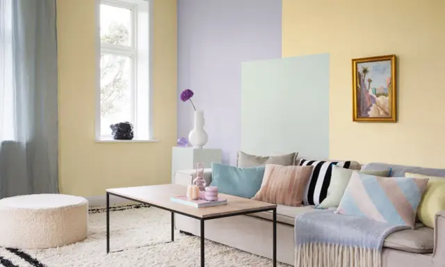 Ett vardagsrum med pastell inredning. Ljusgula, ljuslila och ljusgröna väggar skapar en mjuk atmosfär. En beigefärgad soffa är fylld med kuddar i olika pastellfärger och mönster.