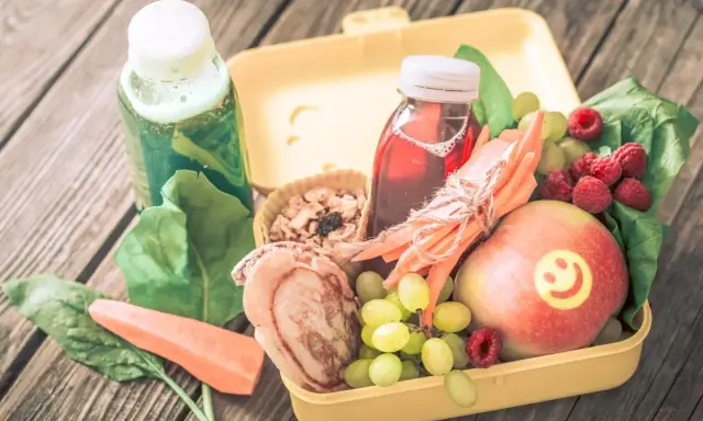 Zdrowa dieta 3 latka: żółty lunchbox z jabłkiem, malinami, winogronami, marchewką, placuszkiem i sokami.