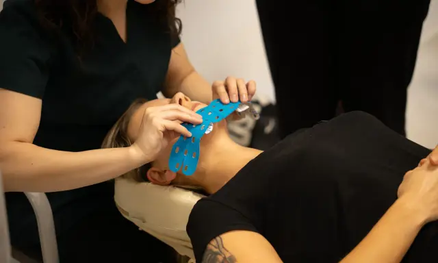 Specjalistka aplikuje niebieskie plastry na twarz pacjentki. Taping twarzy to metoda wspierająca regenerację i poprawiająca wygląd.