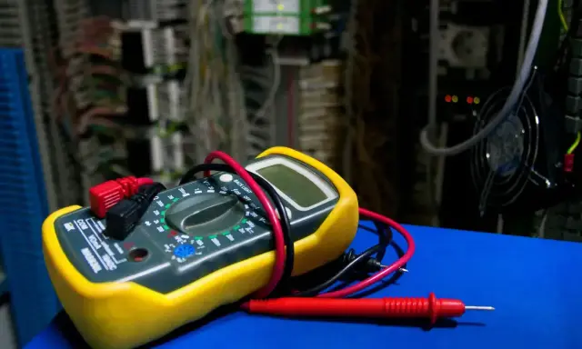 Żółty miernik cyfrowy z czerwonymi i czarnymi przewodami, gotowy do użycia. W tle widać skomplikowaną instalację elektryczną. Dowiedz się, jak używać miernika.