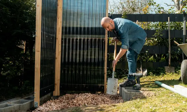 Uomo costruisce una doccia da giardino fai da te, livellando ghiaia con una pala.