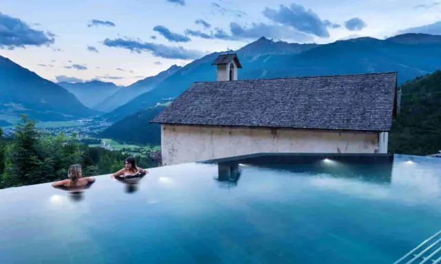 Due donne in una piscina a sfioro ammirano il panorama di Bormio e dintorni, con montagne maestose e un piccolo edificio storico.