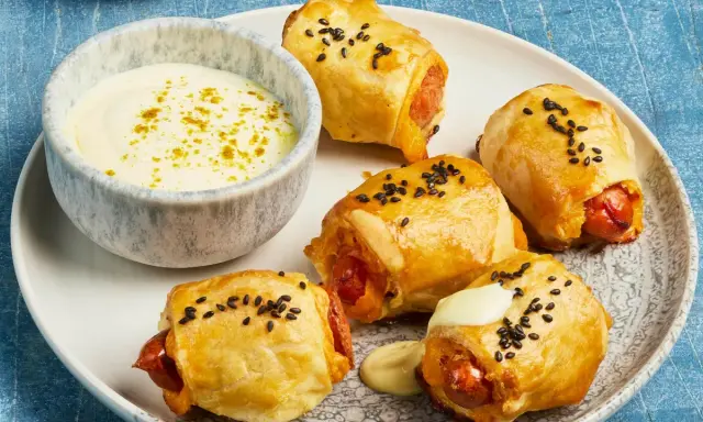 Goldbraune Blätterteigkissen mit Würstchen, bestreut mit schwarzen Sesamkörnern, serviert mit einer cremigen Dip-Sauce.