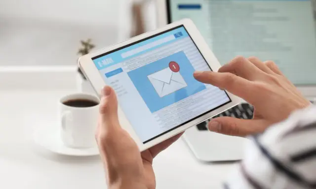 Ręce trzymają tablet z ekranem poczty e-mail, gdzie widać powiadomienie o nowej wiadomości. Może to być początek pisania reklamacji po angielsku.
