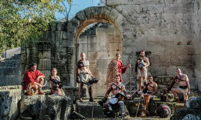Un groupe de guerriers, prêts à combattre, pose devant des ruines antiques. Ces hommes, tels des gladiateurs romains, arborent armures et armes, prêts pour l'arène.