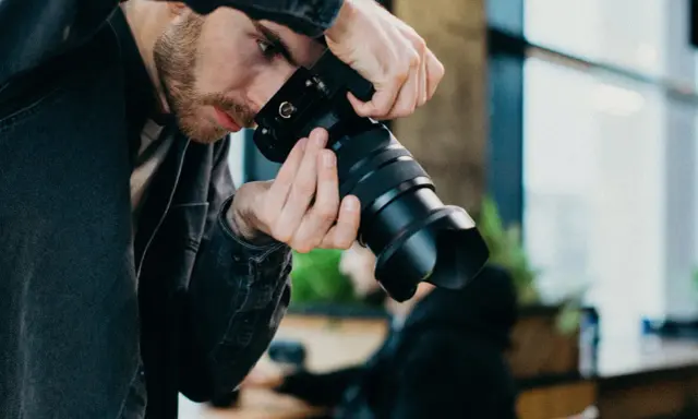 Mężczyzna z brodą w czarnej kurtce trzyma aparat, gotowy do uchwycenia chwili w stylu street photography.